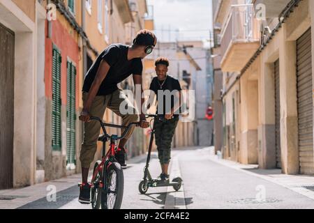 Cheerful young African American man riding electric scooter while black male friend is driving bicycle in street Stock Photo