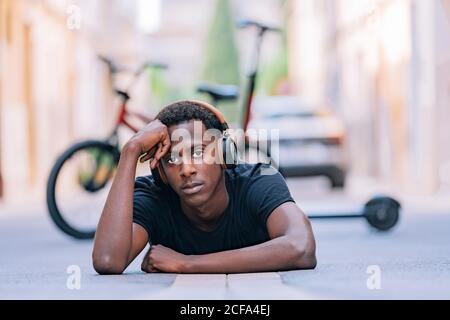 Concentrated youthful African American man wearing headphones listening to music while lying on asphalt road in street Stock Photo