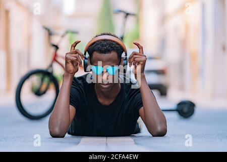 Concentrated youthful African American man in sunglasses wearing headphones and listening to music while lying on asphalt road in street Stock Photo
