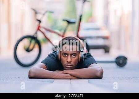 Concentrated youthful African American man wearing headphones listening to music while lying on asphalt road in street Stock Photo