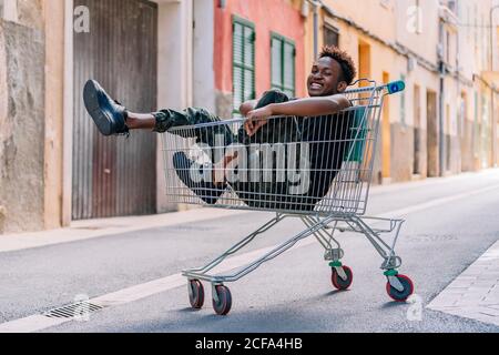 Side view of pensive youthful African American male teenager in casual clothes looking at camera while sitting in metal shopping trolley in street Stock Photo