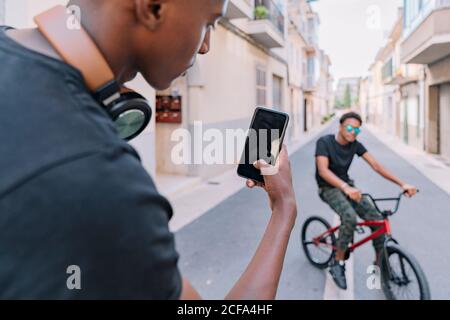 From above crop focused young African American guy taking picture on smartphone of male black friend on bike in street Stock Photo