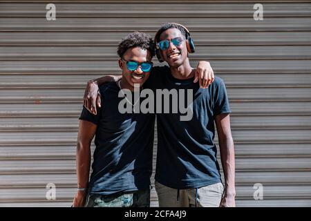 Happy youthful African American male teenagers in sunglasses enjoying pastime together while standing in sunlight in street Stock Photo