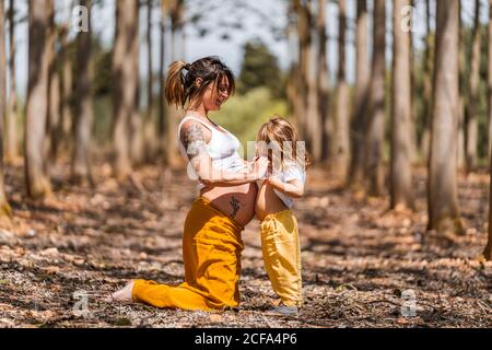 Side view of cheerful barefooted pregnant Woman and little girl in similar clothes holding hands and touching bellies to each other while having fun in autumn forest glade during sunny day Stock Photo
