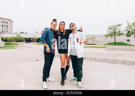 Male and female friends in casual stylish clothing standing with arms around each other while smiling and looking at camera at empty city street Stock Photo