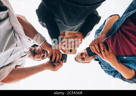 From below of young hipsters in casual wear standing and forming a ring while browsing mobile phones on white background Stock Photo