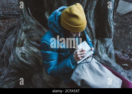 From above of teenage girl in warm active wear with backpack sitting on massive roots of old huge tree and taking notes in small notebook during exciting trekking in autumn forest Stock Photo