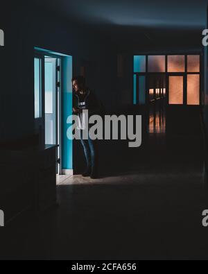 Madagascar - JULY 6, 2019: Curious man in casual clothes standing and leaning on wall by doorway in dark hallway Stock Photo