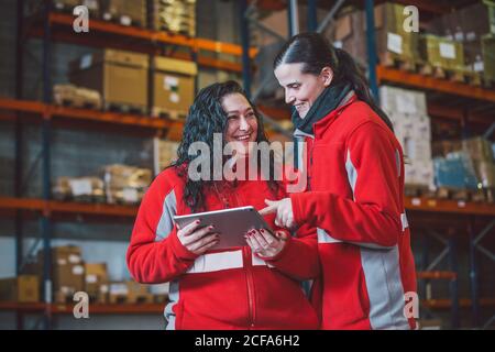 From below of young women in red uniform smiling while using digital tablet during work in modern warehouse Stock Photo