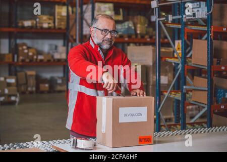 Senior bearded male worker in red uniform opening parcel with paper knife and looking at camera in big storage Stock Photo
