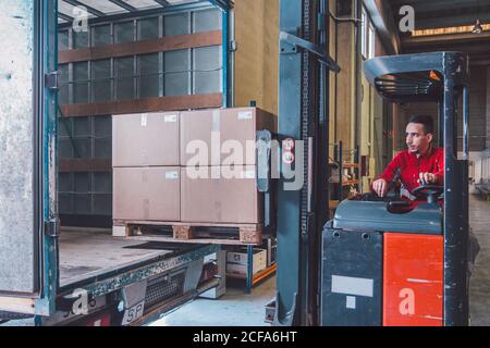 Male driver of forklift truck during shipment packages from big truck in modern warehouse Stock Photo