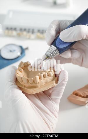 From above of crop anonymous dentist in medical gloves grinding dental prosthesis with professional tool while working in modern laboratory Stock Photo