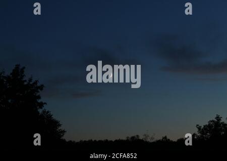 white Crescent a piece of the moon against a blue sky with small clouds and dark silhouettes of trees on the edges as a frame of the night landscape o Stock Photo