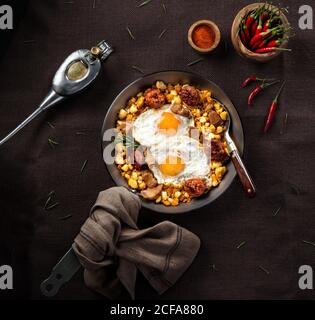 Top view of typical Spanish fried eggs with tortilla crumbs and chorizo sausages with bacon served in frying pan on black table with red pepper Stock Photo