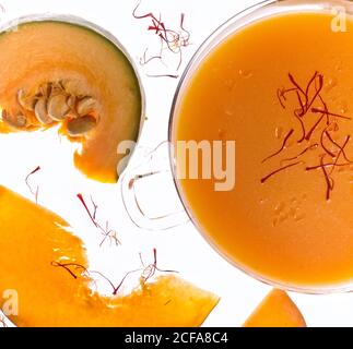 Top view of glass bowl filled with bright aromatic vegetarian pumpkin soup garnished with saffron placed on white table near fresh sliced pumpkin Stock Photo