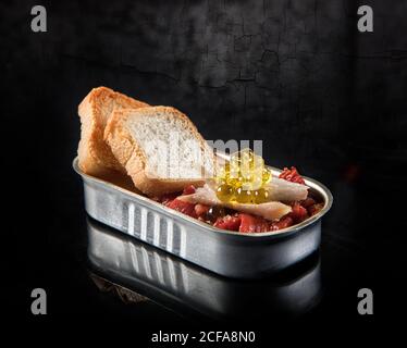 Appetizing roasted red bell peppers with fresh bread and yellow roe with fish meat in tin can on black glossy surface with reflection against gray shabby background in dark room Stock Photo