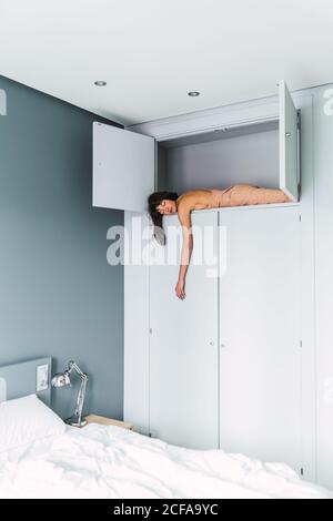 Side view of young Woman sleeping on high shelf of wardrobe near bed in bedroom Stock Photo