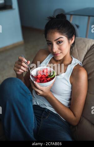 Beautiful brunette Hispanic Woman holding fork and bowl of kiwi and watermelon and sitting on settee Stock Photo