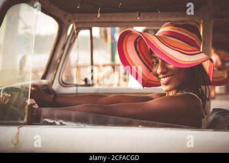 Side view of cheerful young African American female in colorful wide brimmed striped hat sitting behind wheel of vintage van and looking at camera Stock Photo