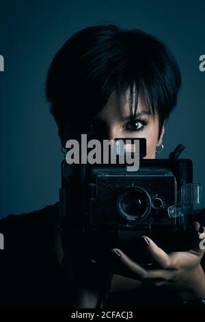 Concentrated youthful dark haired female model looking at camera and photographing with retro camera in studio on blue background Stock Photo