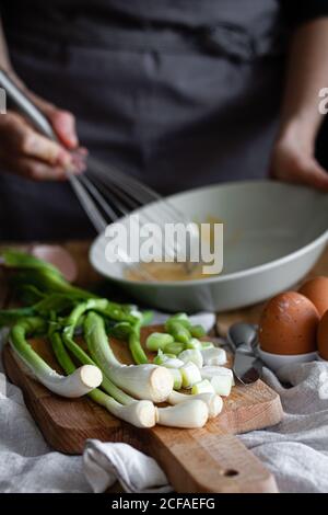 Bunch of fresh scallions and mushrooms placed on cutting board near eggs and poppy seeds against crop housewife mixing ingredients in bowl Stock Photo