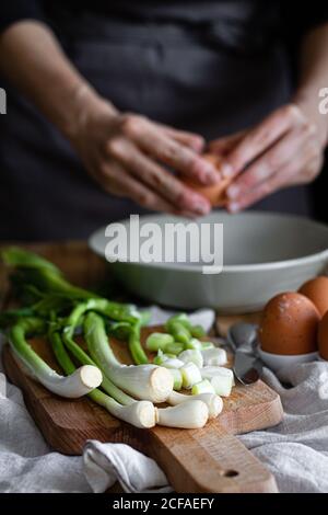 Cropped Woman hands breaking eggs in a bowl with bunch of fresh scallions and mushrooms placed on cutting board near poppy seeds Stock Photo