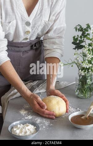 anonymous female baker in apron kneading soft dough with flour on table near apple sauce and bouquet of flowers while cooking pastry Stock Photo