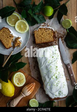 Top view of loaf of delicious pastry with sweet icing placed on wooden table near kitchenware and ripe lemons and limes Stock Photo