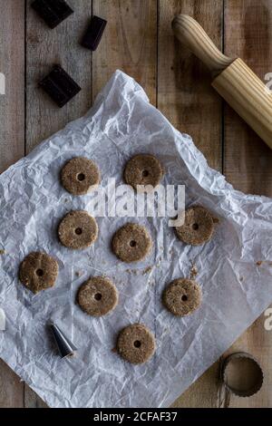 From above chocolate unbaked round cookies on white baking paper with chocolate metal cookie molds and rolling pin on wooden table Stock Photo