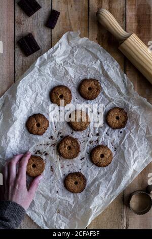 From above anonymous person hand holding chocolate unbaked round cookies on white baking paper with chocolate metal cookie molds and rolling pin on wooden table Stock Photo