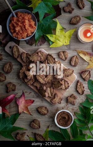 Top view of tasty biscuits and autumn leaves placed on wooden table near cocoa powder and sweet paste Stock Photo