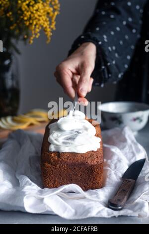 Crop hand of housewife decorating delicious homemade lemon and poppy seeds sponge cake with whipped cream at table with bouquet of mimosa flowers in background Stock Photo