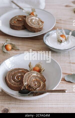 From above of sliced delicious homemade sweet roll cake with whipped cream and Physalis dried flowers served on plate on wooden table with ingredients Stock Photo