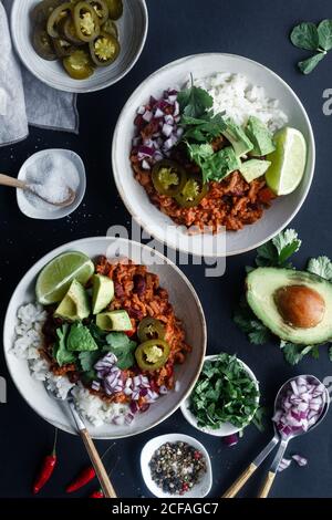 Top view of appetizing traditional Mexican chili with meat and white rice served in bowls with avocado and lime with chopped onion and marinated green chili pepper on black background with ingredients Stock Photo