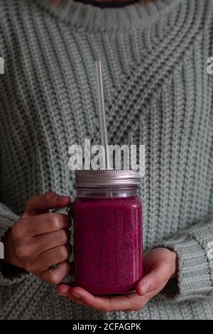 Crop person in casual knitted sweater holding transparent jar with metal lid and plastic straw of tasty vitamin pink beverage of sweet fresh fruits and berries while having healthy appetite breakfast Stock Photo