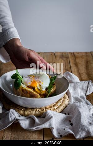 Crop anonymous hand holding dish with fried egg on potato on wooden table with fried mushrooms grated cheese and herbs Stock Photo