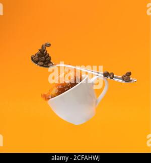 Falling white cup with splashing hot coffee and teaspoon with beans of coffee on yellow background Stock Photo