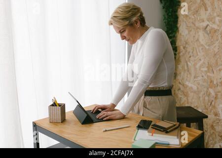 Focused blond adult Woman standing at wooden table with stationery typing on portable keypad of tablet against light window Stock Photo
