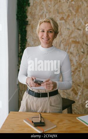 Happy focused adult businessWoman looking at camera while using mobile phone near notepad on wooden table in light office Stock Photo