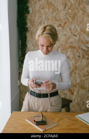 Focused adult businessWoman looking at camera while using mobile phone near notepad on wooden table in light office Stock Photo