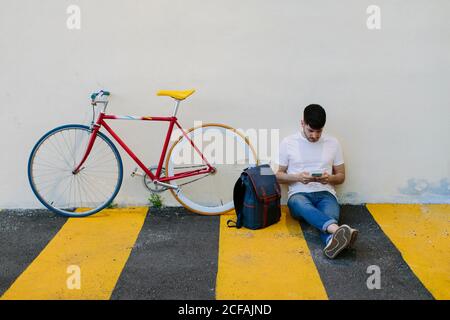 Front view of a caucasian man with a smartphone sitting on the floor with a colorful bike in the street. Stock Photo