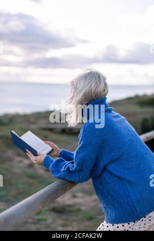 Side view of unrecognizable young female in blue sweater leaning on wooden fence and enjoying favorite poetry while spending time at seaside Stock Photo