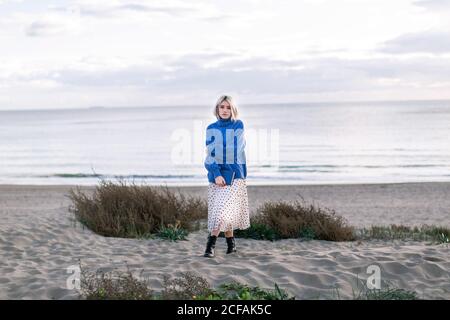Thoughtful young Woman with blonde hair in warm blue sweater holding book and looking at camera while standing against ocean sandy background Stock Photo