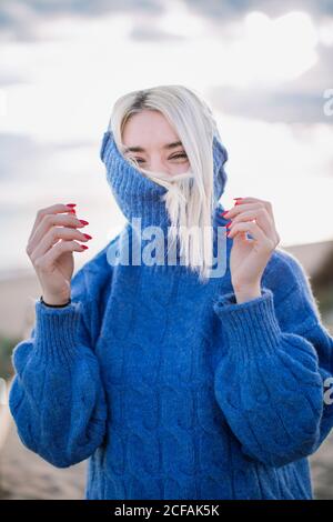 Joyful cheerful young blonde female in blue knitted sweater looking at camera and laughing while standing against blurred background of sea coast Stock Photo