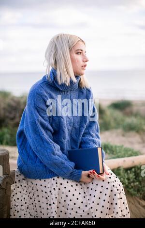 Thoughtful young blonde female in warm blue sweater sitting on wooden fence holding book while spending spring day in countryside looking away Stock Photo