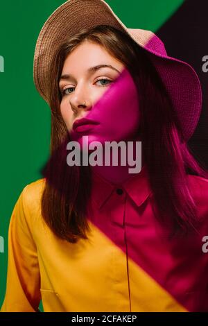 Young female model in stylish hat with red shadow on face and shoulder looking at camera against green background
