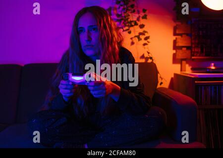 Young Woman playing video games in her house during Corona Times. She is in a living room with colorful lights at night. Stock Photo