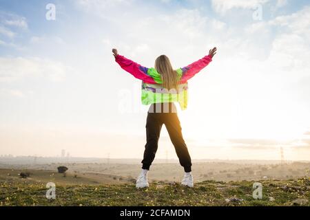 Back view of contemporary blond haired young Woman in colorful jacket raising hands with remote land on background Stock Photo