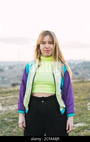 Contemporary cheerful blond haired young Woman in colorful jacket and green neon top looking at camera with remote land on background Stock Photo