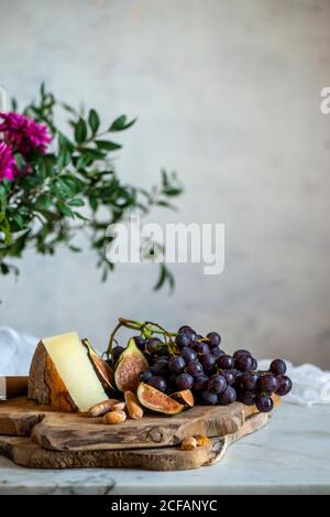 Sliced ripe figs and dark blue grapes next to piece of cheese on wooden cutting boards near bouquet of pink flowers among green leaves against blurred gray wall Stock Photo